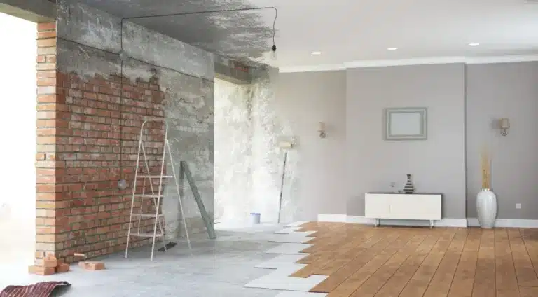 A living space being remodeled. A ladder sits beside a brick wall on a concrete floor partially covered in wood flooring