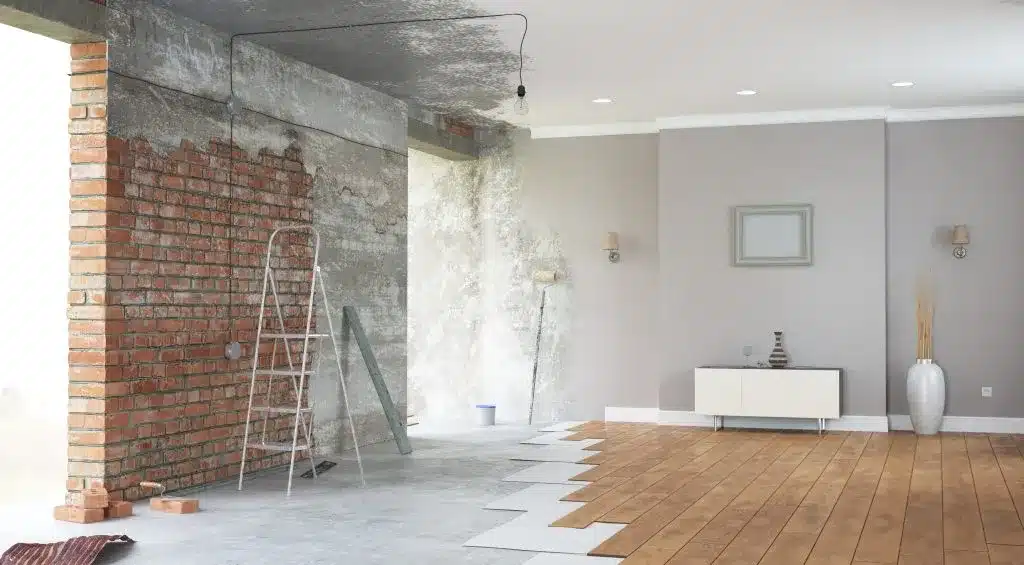 A living space being remodeled. A ladder sits beside a brick wall on a concrete floor partially covered in wood flooring